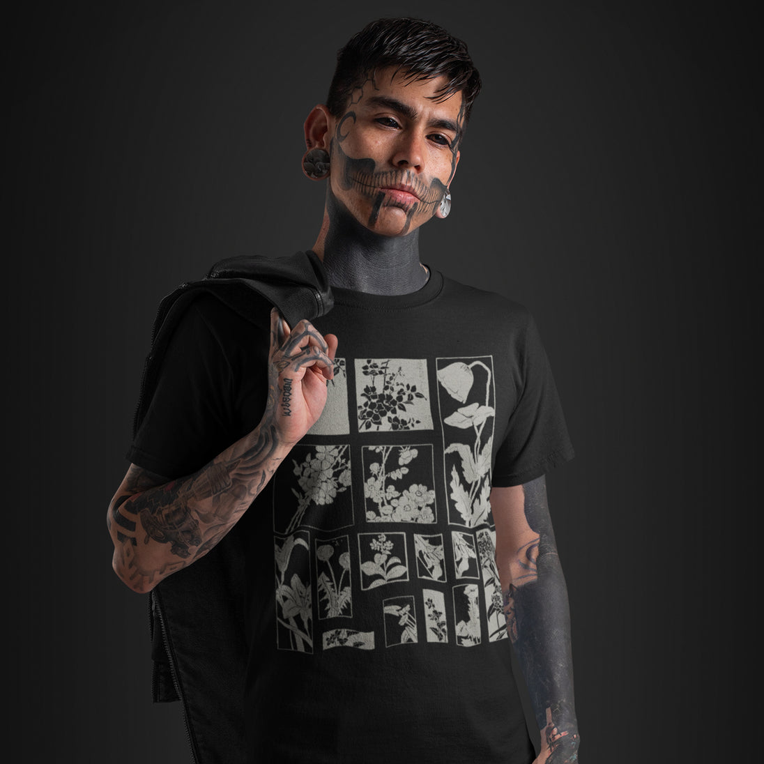 A man with face tattoos and gray face paint wears a black graphic t-shirt featuring a white grid pattern of floral illustrations. He is posed against a dark studio background.
