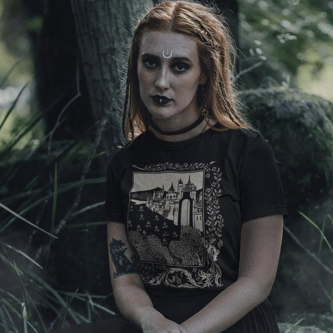Woman with a tattooed arm wearing a black Medieval Castle T-Shirt, sitting in a forest.