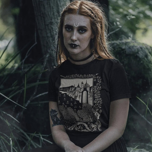 Woman with a tattooed arm wearing a black Medieval Castle T-Shirt, sitting in a forest.