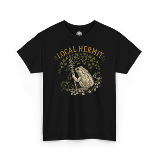 A black short-sleeve crewneck t-shirt featuring a vintage-style graphic of a toad holding a staff, surrounded by flowers and leaves, with the text 'LOCAL HERMIT' printed above in a warm tan color.