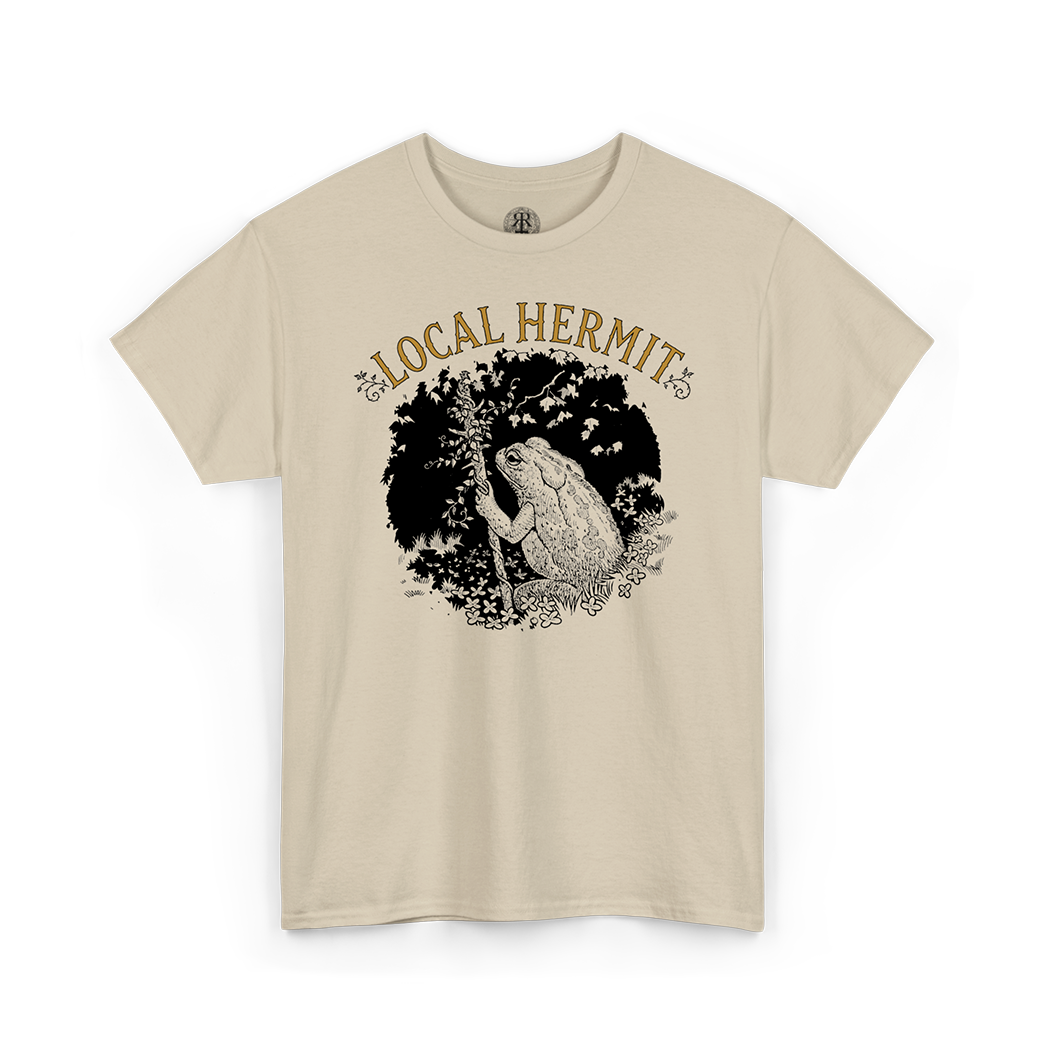 A beige short-sleeve crewneck t-shirt featuring a graphic print of a toad in a black circle with the text 'LOCAL HERMIT' in tan above it, displayed on a clean white background.