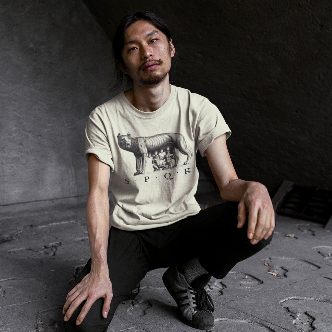 Man wearing a beige t-shirt with a SPQR Capitoline Wolf graphic design, sitting on the ground in a dark room.