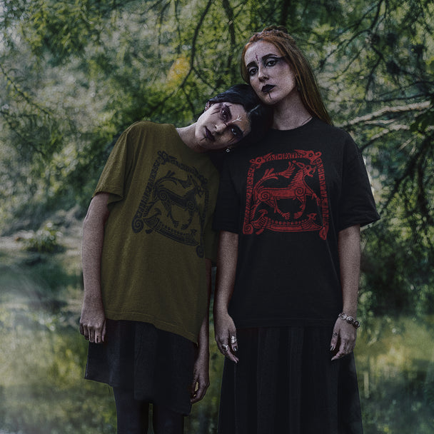 Two models posing outdoors in gothic-style graphic t-shirts. The model on the left wears an olive green oversized t-shirt with a black dragon graphic, and the model on the right wears a black crewneck t-shirt with a red Viking horned animal graphic.