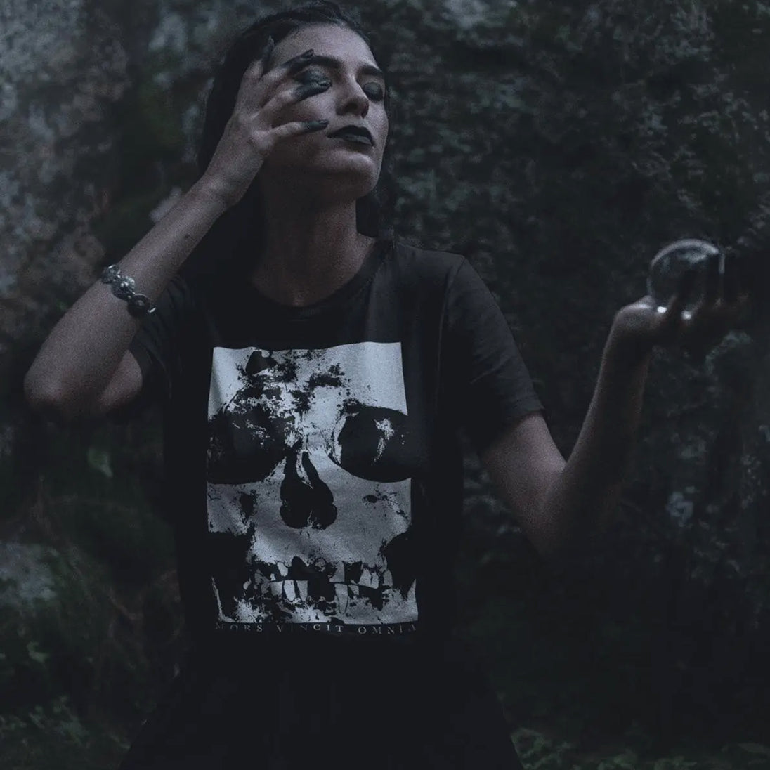 Person wearing a black t-shirt with a graphic design, holding a stone in a dark forest setting.