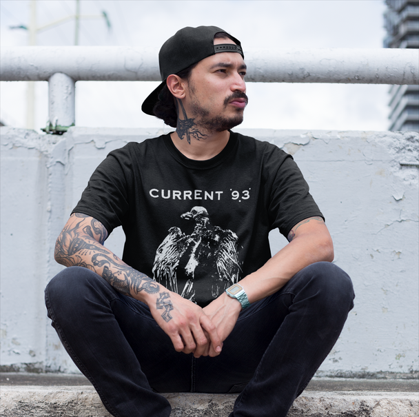 A man wearing a black t-shirt with a white graphic print that reads 'CURRENT '93' and a black baseball cap sits cross-legged against a weathered concrete wall.