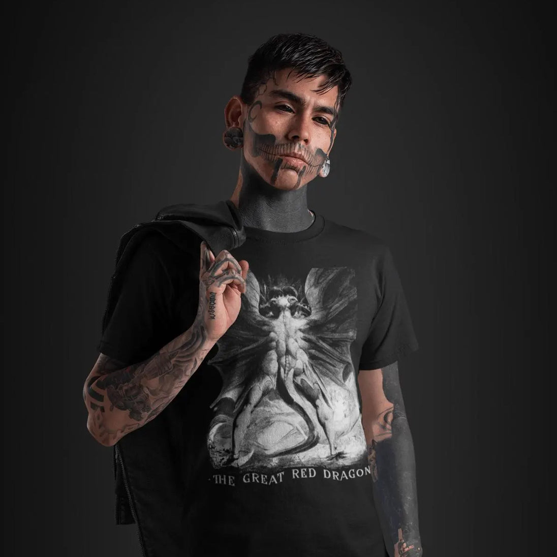 A tattooed man with face paint wears a black graphic t-shirt featuring a grayscale illustration of a winged creature and the text 'THE GREAT RED DRAGON'. He is posed against a dark studio background, holding a black garment over his shoulder.