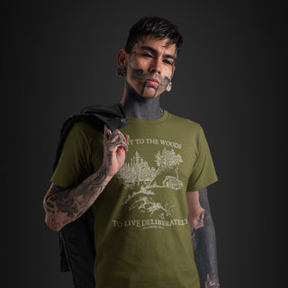 A man with face tattoos and ear piercings wearing an olive green graphic t-shirt with a white sketch of trees and the text 'I WENT TO THE WOODS TO LIVE DELIBERATELY'. He has a black jacket slouched over his shoulder against a dark studio background.