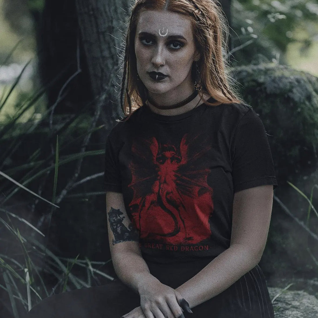 A woman with gothic makeup and a crescent moon forehead sticker sits outdoors in a forest, wearing a black t-shirt with a red graphic of a dragon and the text 'GREAT RED DRAGON'.