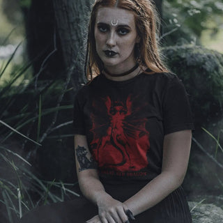 A woman with gothic makeup and a crescent moon forehead sticker sits outdoors in a forest, wearing a black t-shirt with a red graphic of a dragon and the text 'GREAT RED DRAGON'.