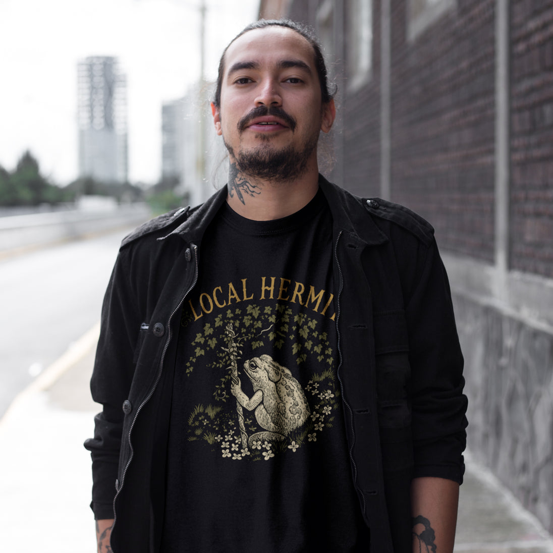 A man wearing a black graphic t-shirt with a frog design and 'LOCAL HERM' text, layered under a black jacket, standing outdoors next to a brick wall.