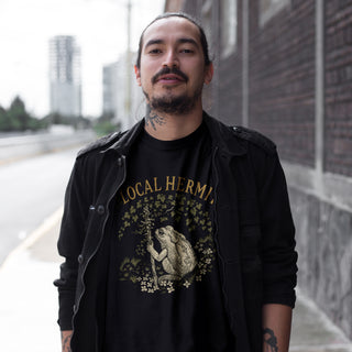 A man wearing a black graphic t-shirt with a frog design and 'LOCAL HERM' text, layered under a black jacket, standing outdoors next to a brick wall.