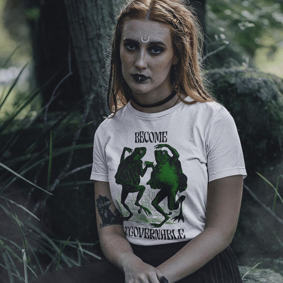 A woman with red hair and gothic makeup sits outdoors in a forest setting, wearing a white graphic t-shirt with a green frog design and the text 'BECOME UNGOVERNABLE'.