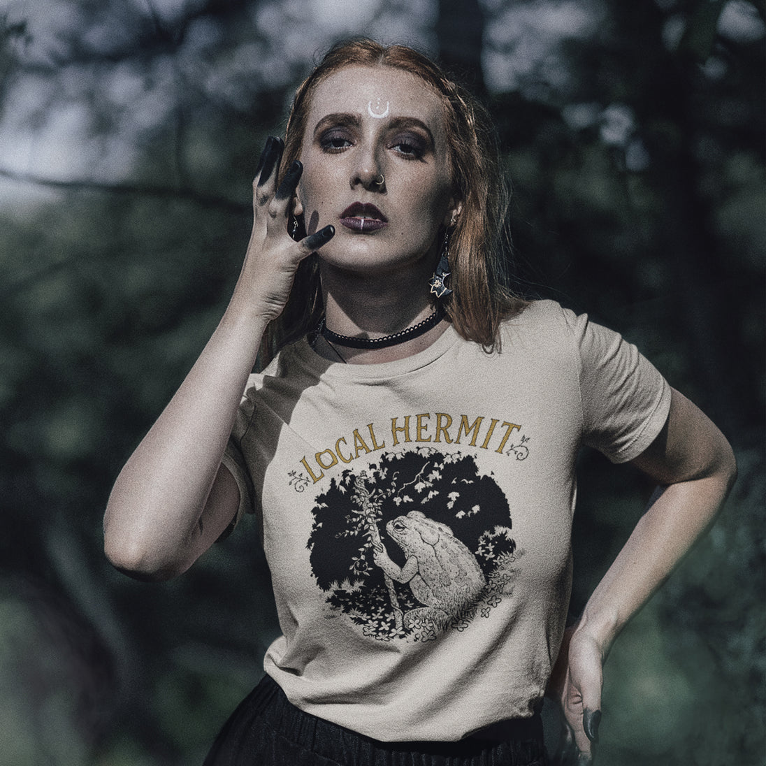 A woman posing outdoors in a beige graphic t-shirt with 'LOCAL HERMIT' and a hermit crab design, set against a dark, blurred forest background.