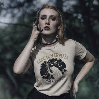A woman posing outdoors in a beige graphic t-shirt with 'LOCAL HERMIT' and a hermit crab design, set against a dark, blurred forest background.