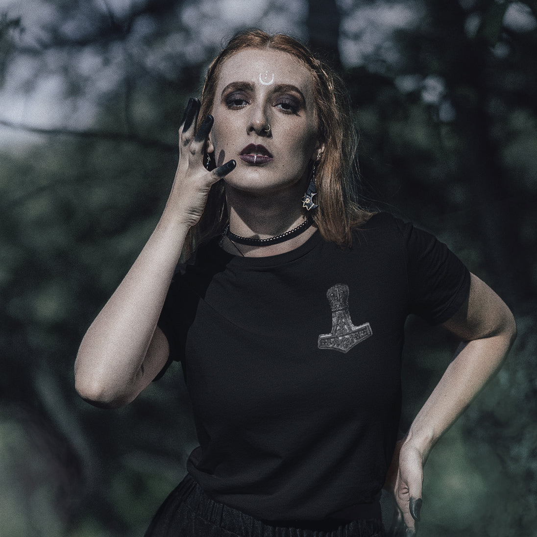 A woman with red hair and gothic makeup wearing a black t-shirt with a silver Mjolnir graphic, posing outdoors against a dark, blurred forest background.