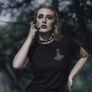 A woman with red hair and gothic makeup wearing a black t-shirt with a silver Mjolnir graphic, posing outdoors against a dark, blurred forest background.