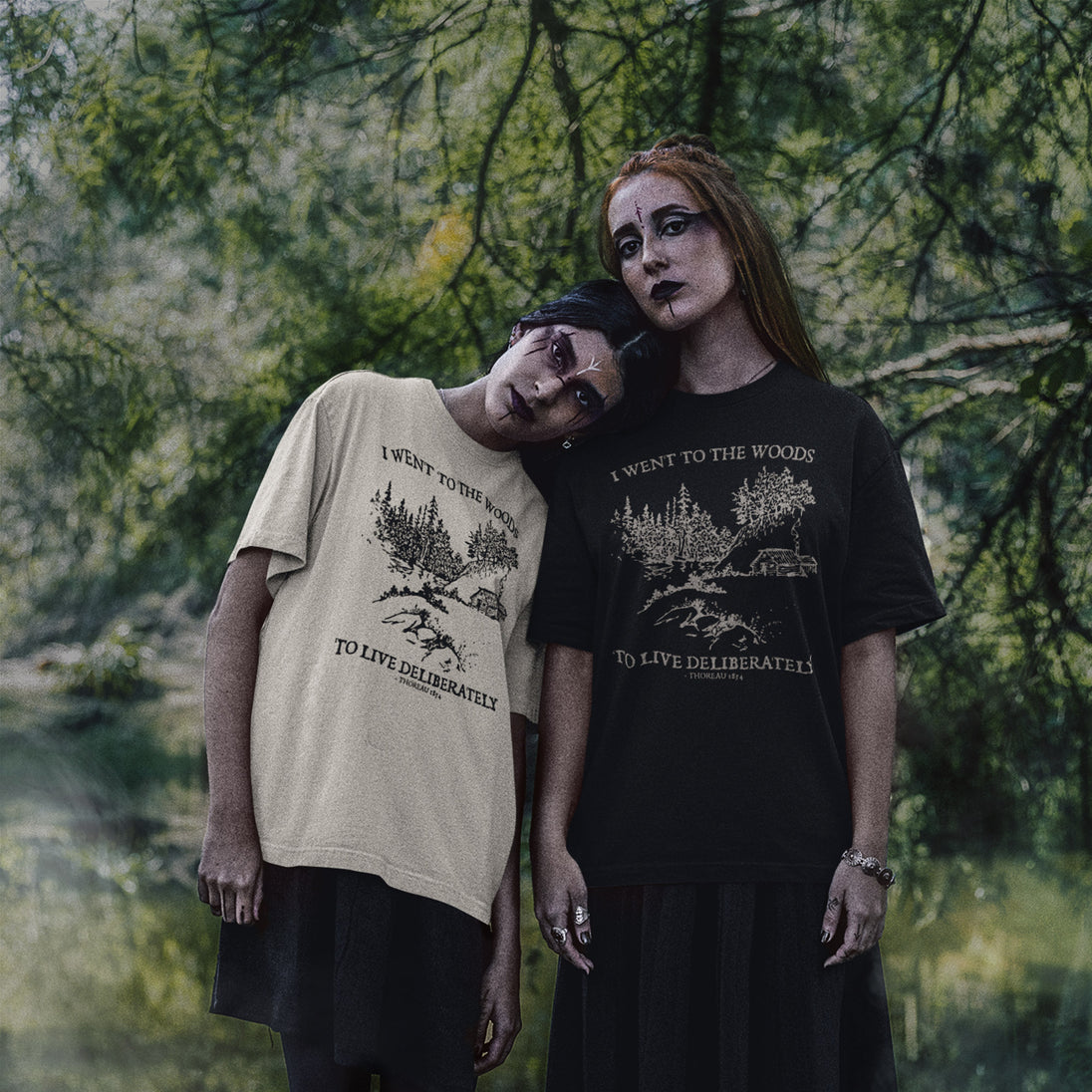Two models posing outdoors in forest surroundings, wearing graphic t-shirts. One model wears a beige t-shirt with a black graphic and text, and the other wears a black t-shirt with a beige graphic and text.