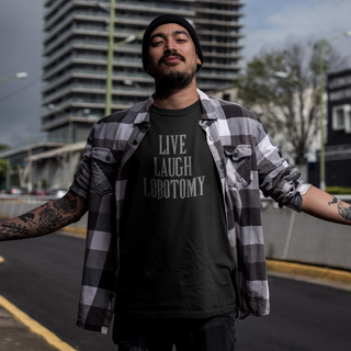 Man wearing a black t-shirt with 'LIVE LAUGH LOBOTOMY' text, plaid shirt, and tattoos outdoors.