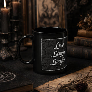 A black ceramic mug with a gothic-style white graphic that reads 'Live Laugh Lucifer' surrounded by a house illustration, set on a dark tabletop with candles, old books, and parchment in the background.