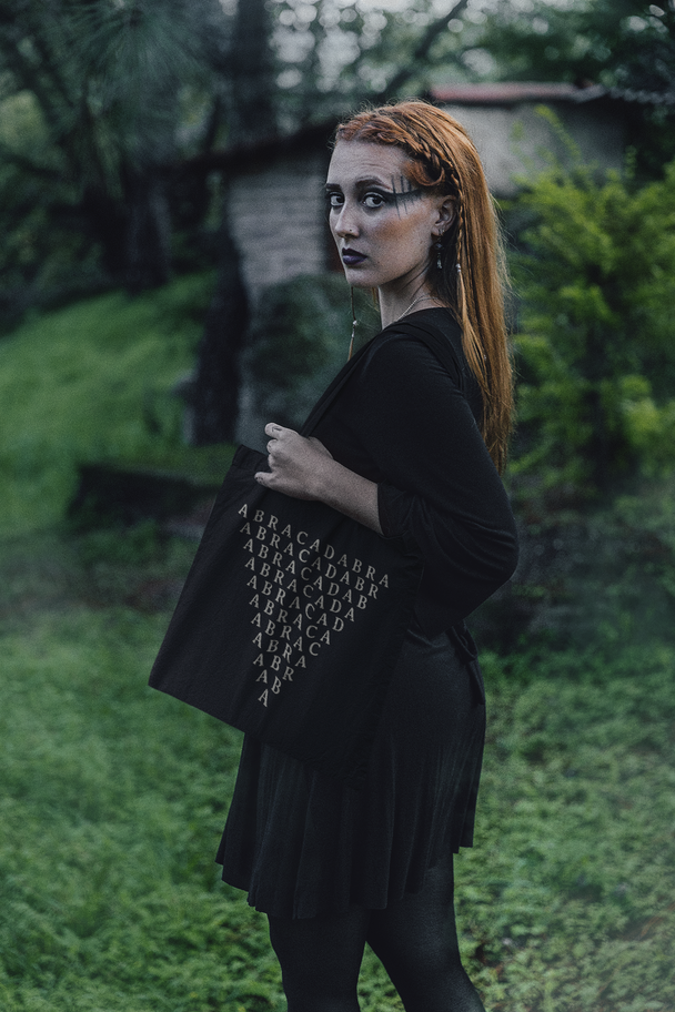 Woman wearing a black outfit with a black tote bag with symbol of Abracadabra inverted triangle