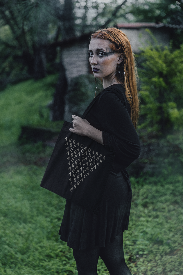 Woman wearing a black outfit with a black tote bag with symbol of Abracadabra inverted triangle