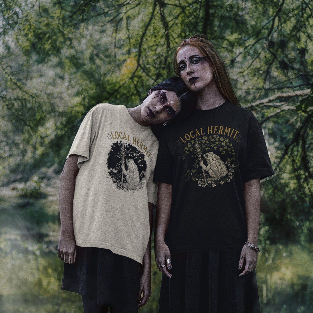 Two models posing outdoors in front of a forest background, wearing graphic t-shirts. The model on the left wears a beige t-shirt with a black graphic and text reading 'LOCAL HERMIT', while the model on the right wears a black t-shirt with a gold graphic and text reading 'LOCAL HERMIT'.