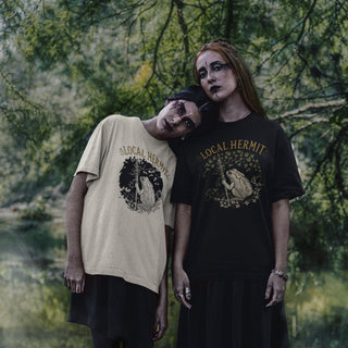 Two models posing outdoors in front of a forest background, wearing graphic t-shirts. The model on the left wears a beige t-shirt with a black graphic and text reading 'LOCAL HERMIT', while the model on the right wears a black t-shirt with a gold graphic and text reading 'LOCAL HERMIT'.