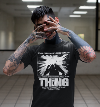 A man with extensive arm and hand tattoos poses in a tiled indoor space, wearing a black graphic t-shirt featuring a white sci-fi design inspired by John Carpenter's 'The Thing'.