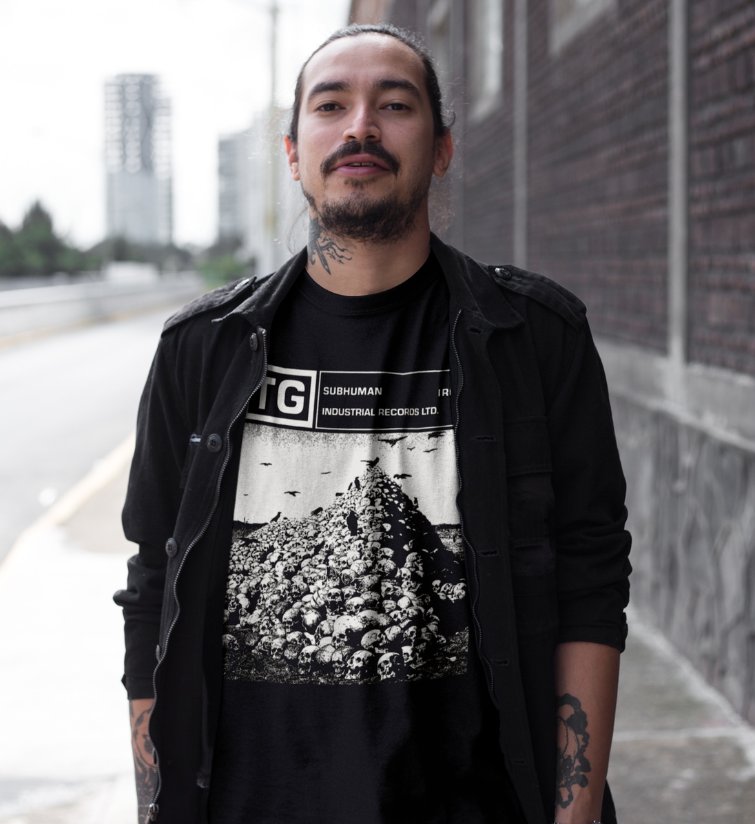 A man wearing a black graphic t-shirt with a skull pile design and 'SUBHUMAN INDUSTRIAL APPAREL LTD.' text, layered under a black jacket, standing outdoors against a brick wall.
