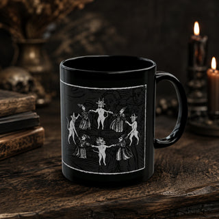 A black ceramic mug with a white gothic illustration of dancing figures, displayed on a rustic wooden table with candles and books in the background.