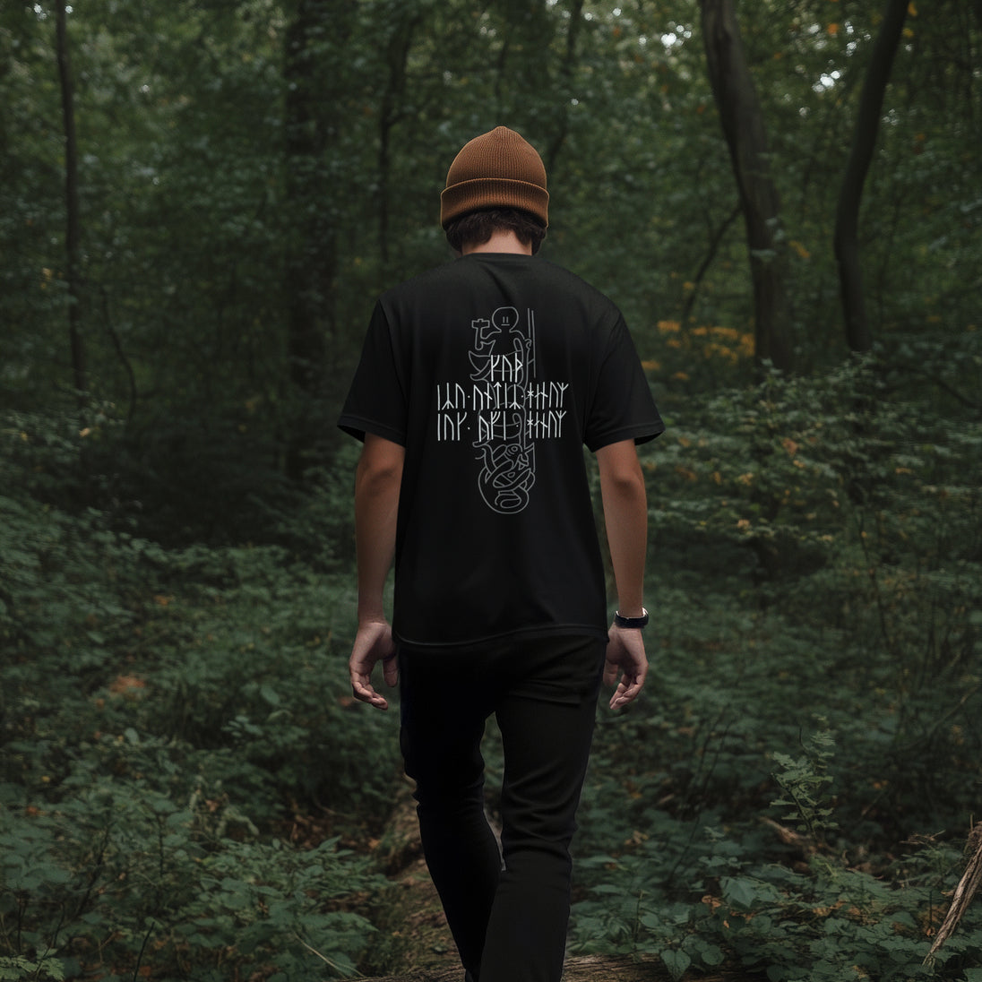 A man viewed from behind walking on a forest path, wearing a black t-shirt with a white graphic print on the back, black pants, and a brown beanie.