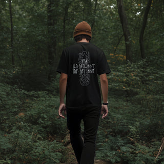 A man viewed from behind walking on a forest path, wearing a black t-shirt with a white graphic print on the back, black pants, and a brown beanie.